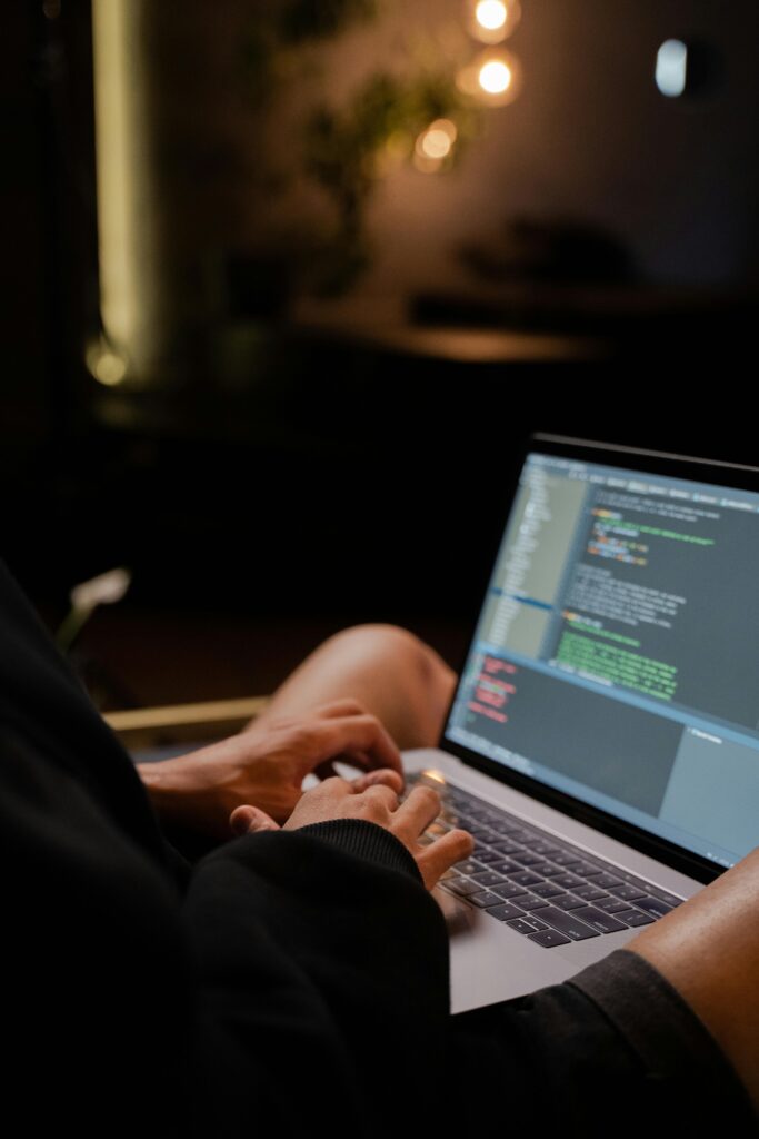 Focused hands typing code on a laptop in a dimly lit room, showcasing programming activities.